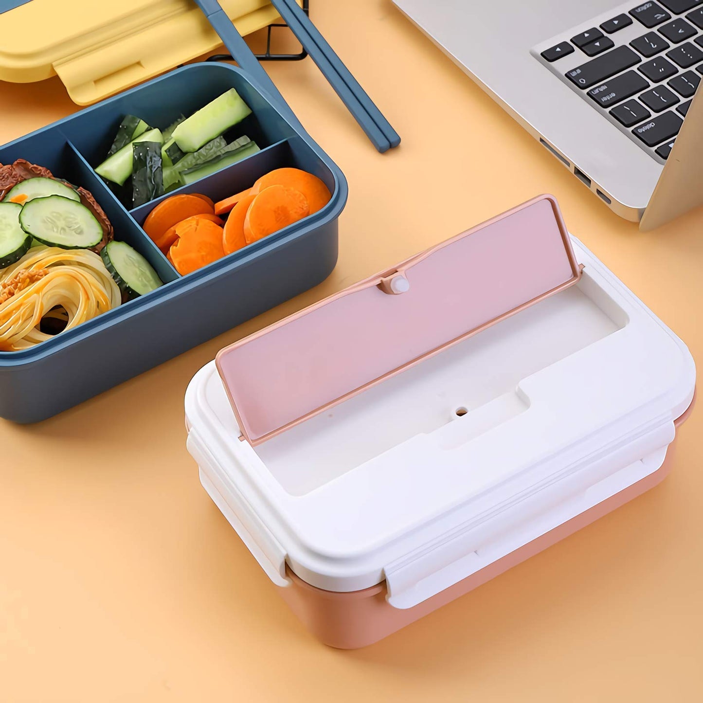 Pink and white lunch box with compartments on a yellow surface, next to a blue bento box with food and a laptop.
